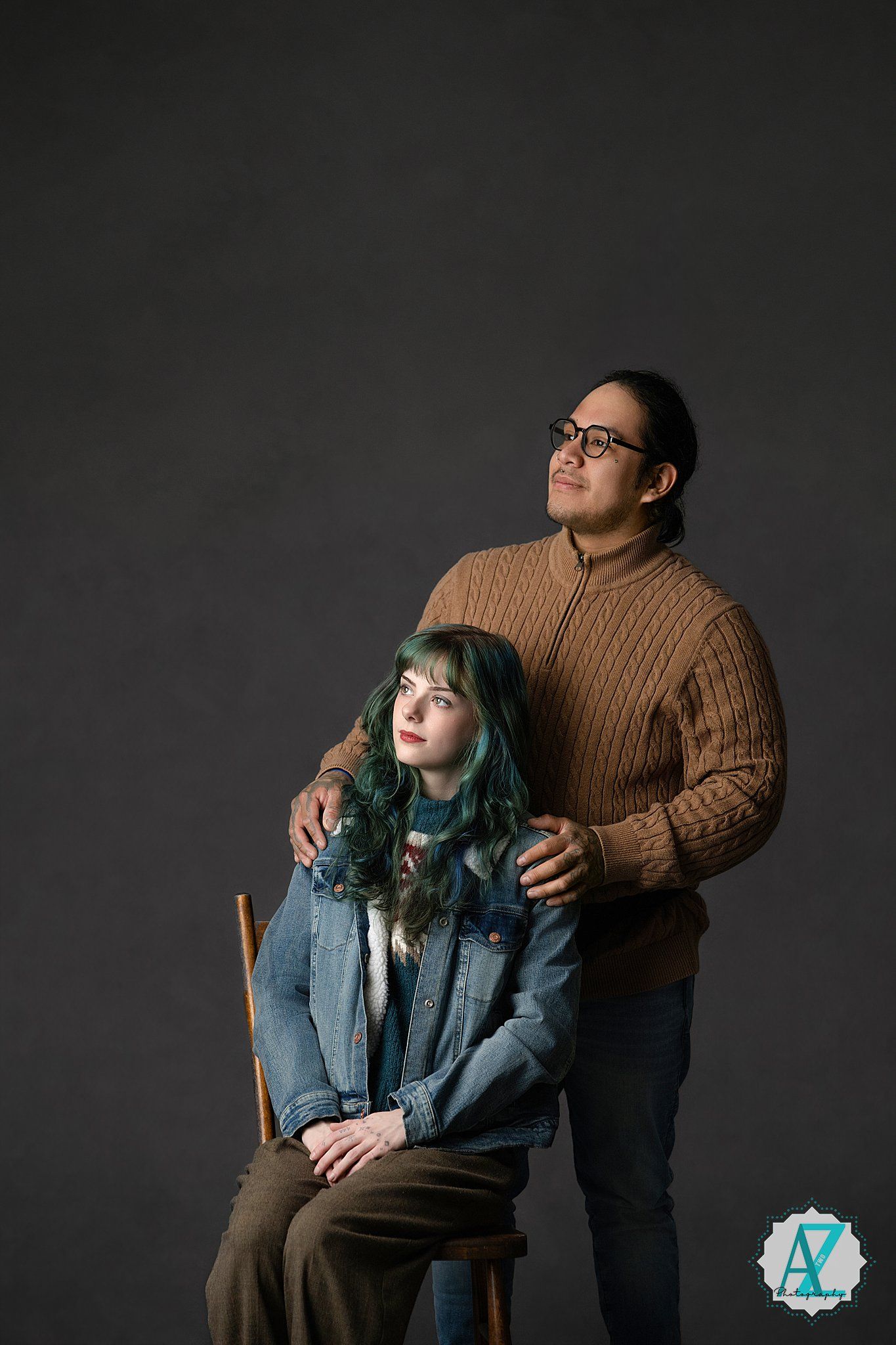 Two people pose together against dark background, one sitting while other stands in brown sweater.