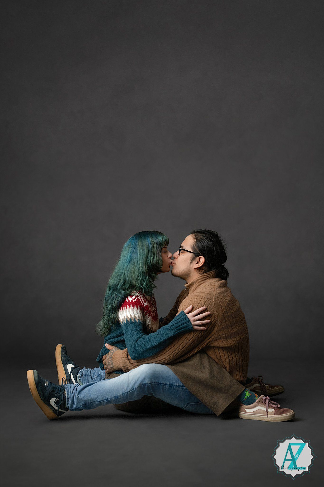 Two people in casual attire sitting intimately on dark floor against gray background sharing romantic moment.