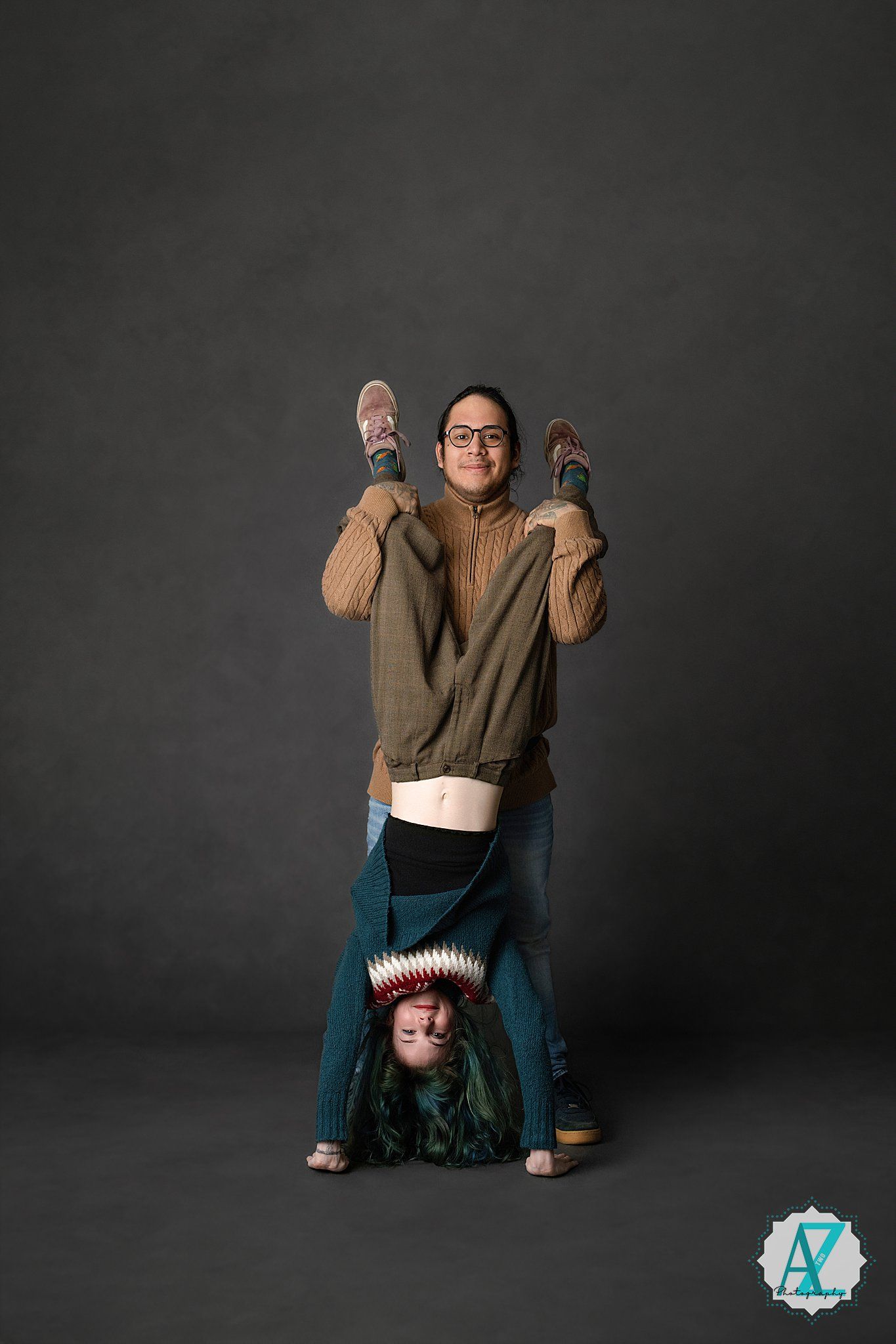 Two people pose playfully against a dark background, with one holding shoes while the other does a handstand.