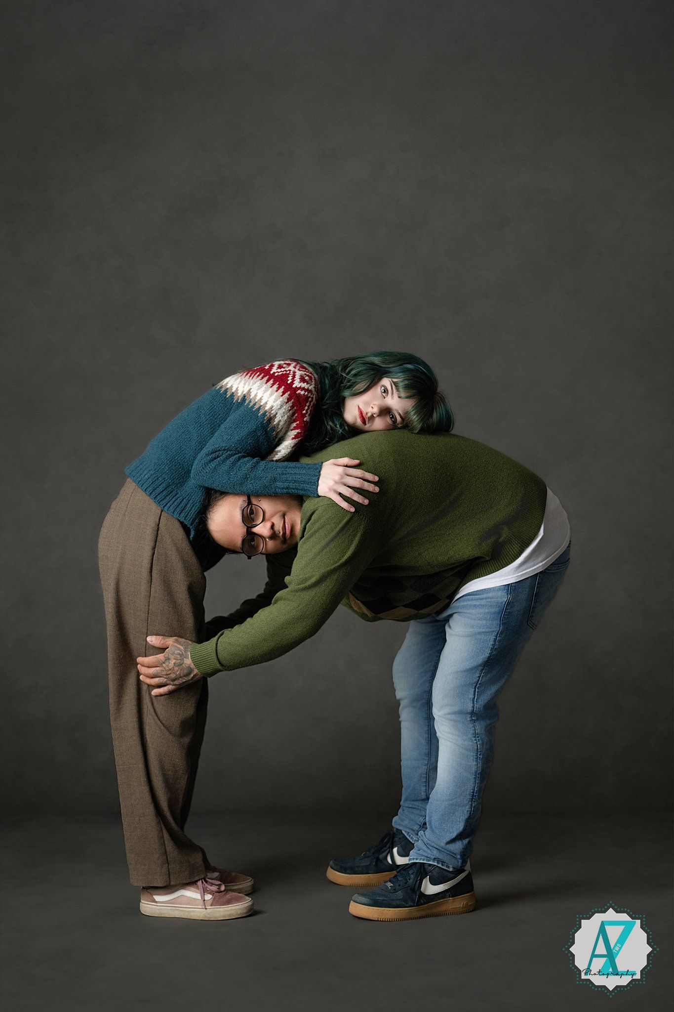 Two people in warm sweaters create a playful curved pose together against a dark gray studio background.