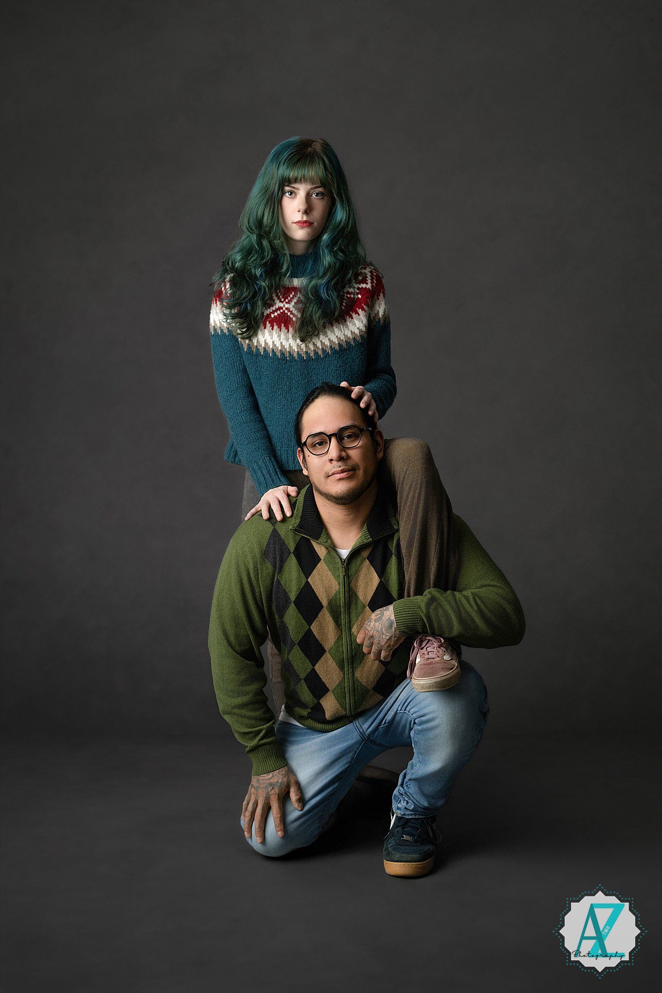 Two people in winter sweaters pose together against a dark background in a studio portrait.