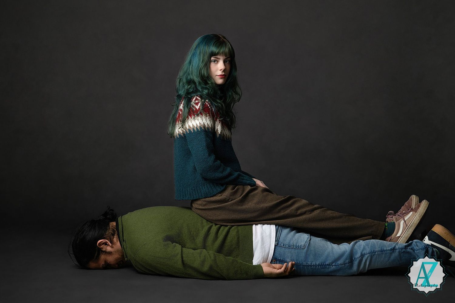 A person in a teal sweater stands over someone lying on the ground in a dramatic studio portrait setup.