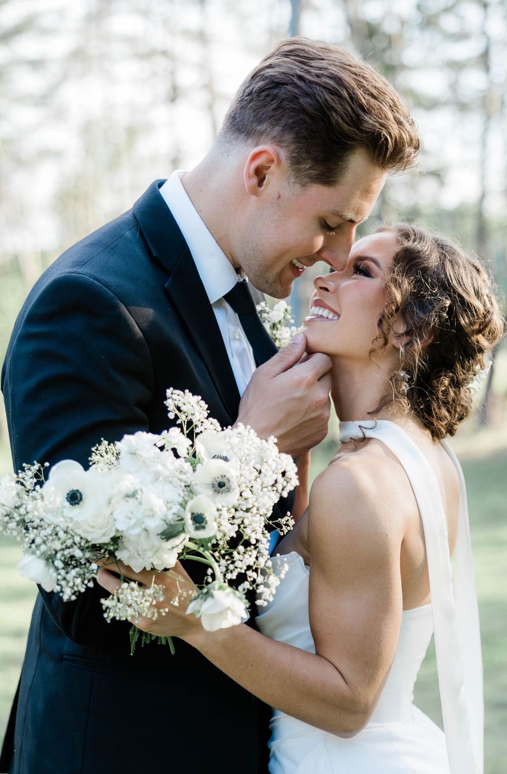 A romantic wedding moment as the couple shares an intimate laugh while holding a white floral bouquet outdoors.