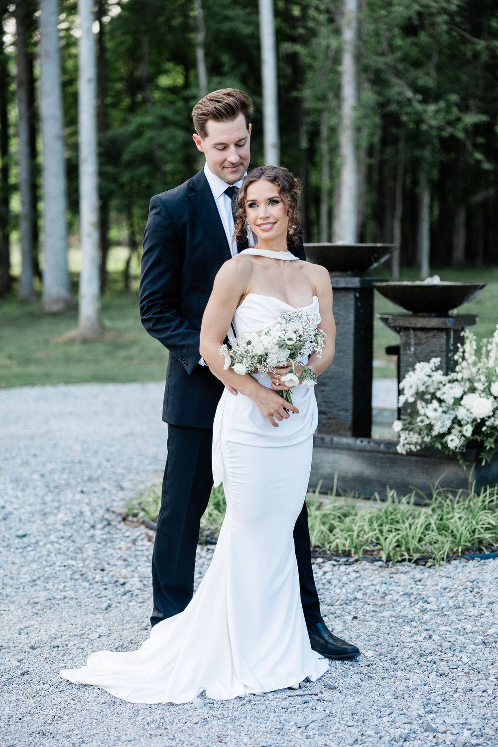 Newlyweds pose together on a gravel path with a forest backdrop, bride in fitted white gown holding bouquet.