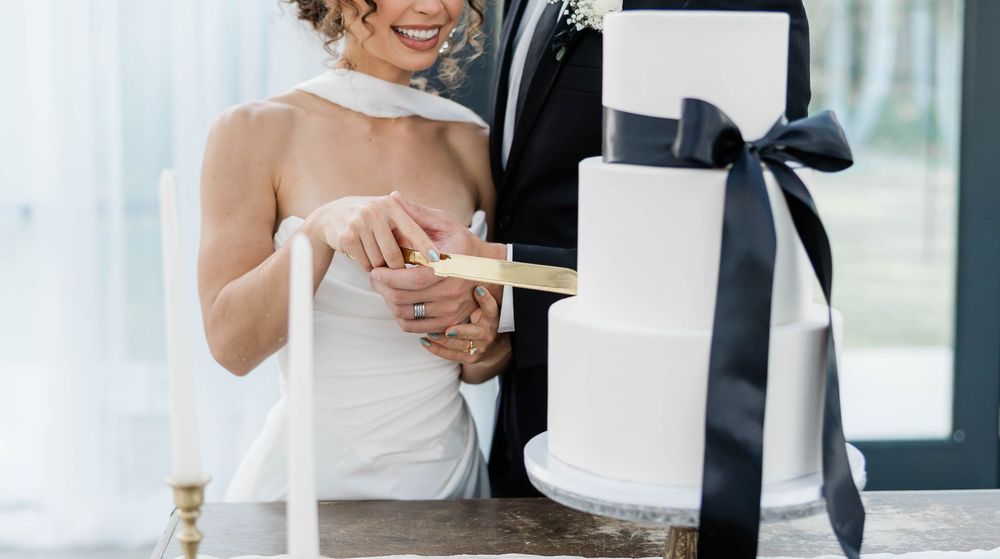 Modern white wedding cake with navy blue ribbon being cut during reception celebration.