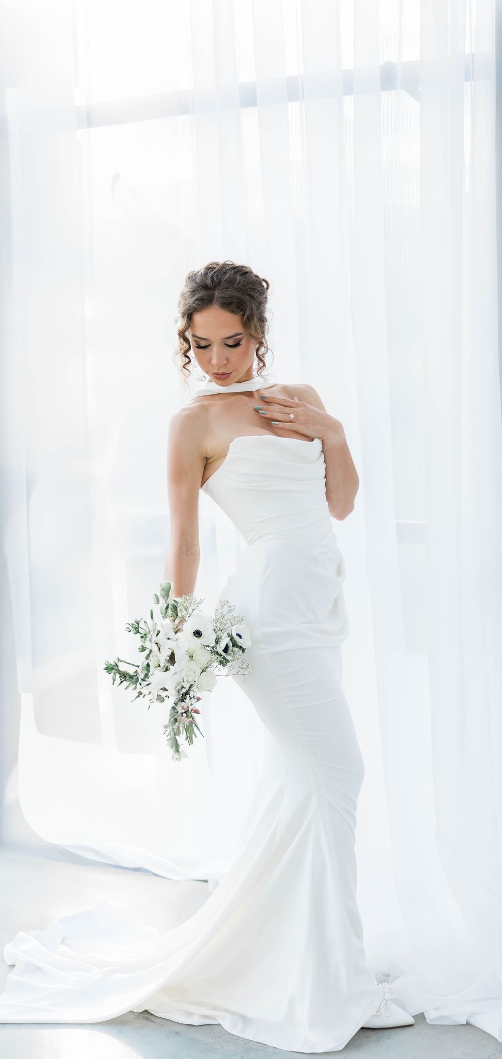 Elegant bride in fitted white gown holding delicate white bouquet against sheer curtains.