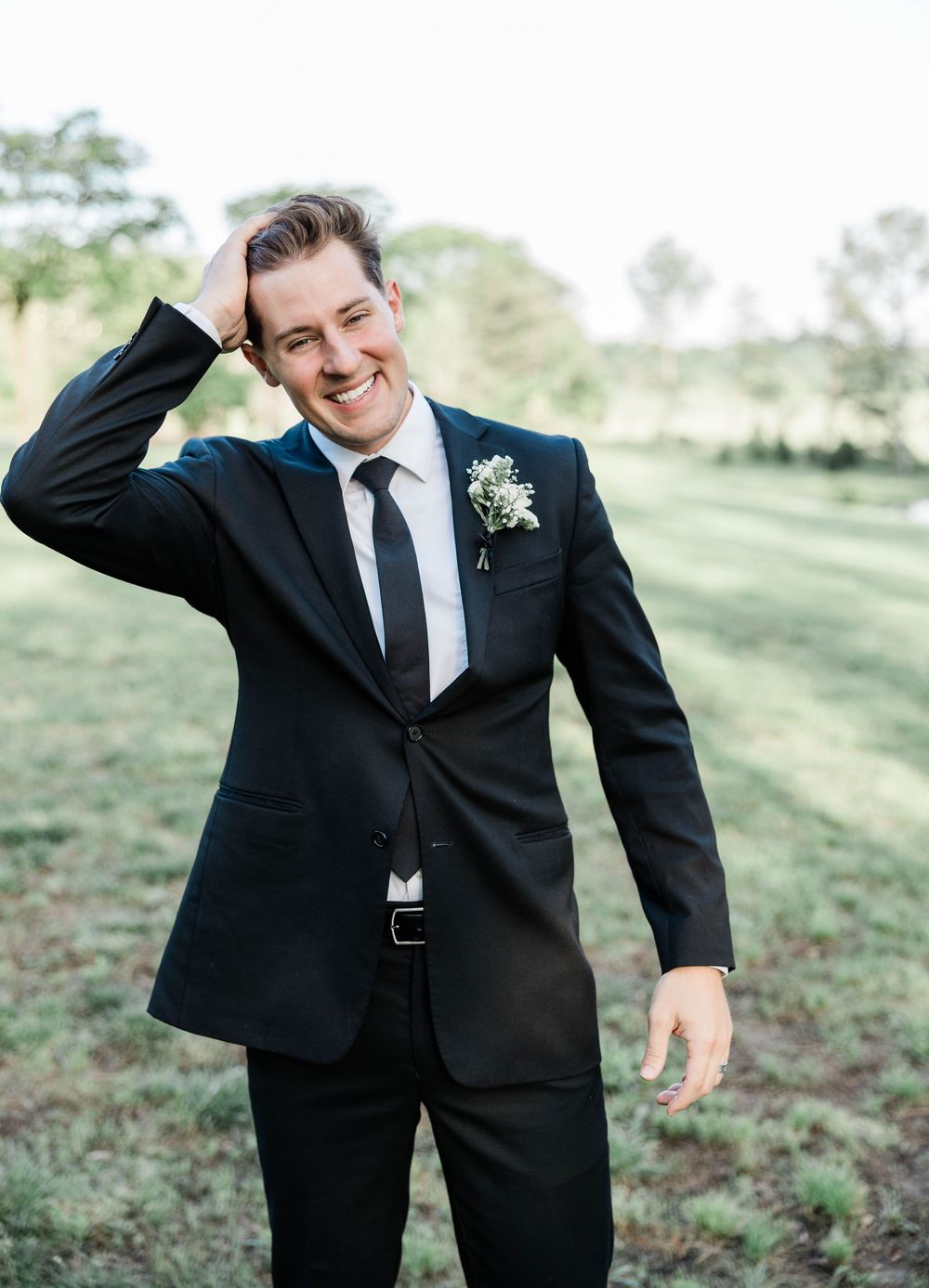 Groom in classic black suit laughing outdoors with boutonniere on lapel.