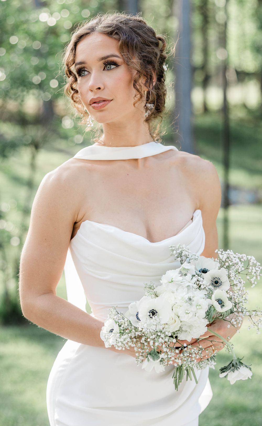 Elegant bride in fitted white gown holding delicate white and green bouquet outdoors.