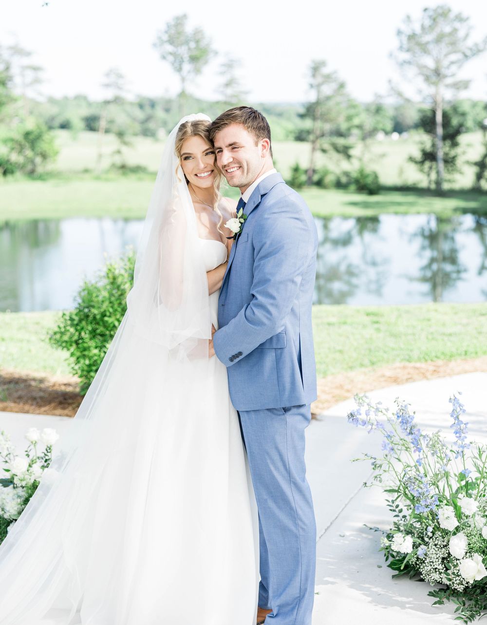 Bright outdoor wedding portrait with lake backdrop and delicate floral arrangements.
