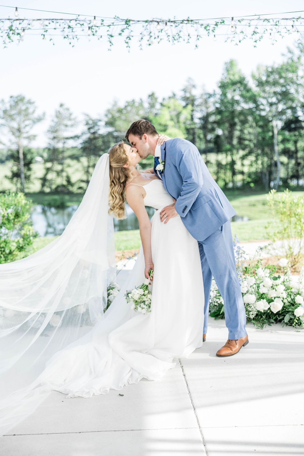 Newlyweds share a romantic dip kiss outdoors with flowing veil and light blue suit against nature backdrop.