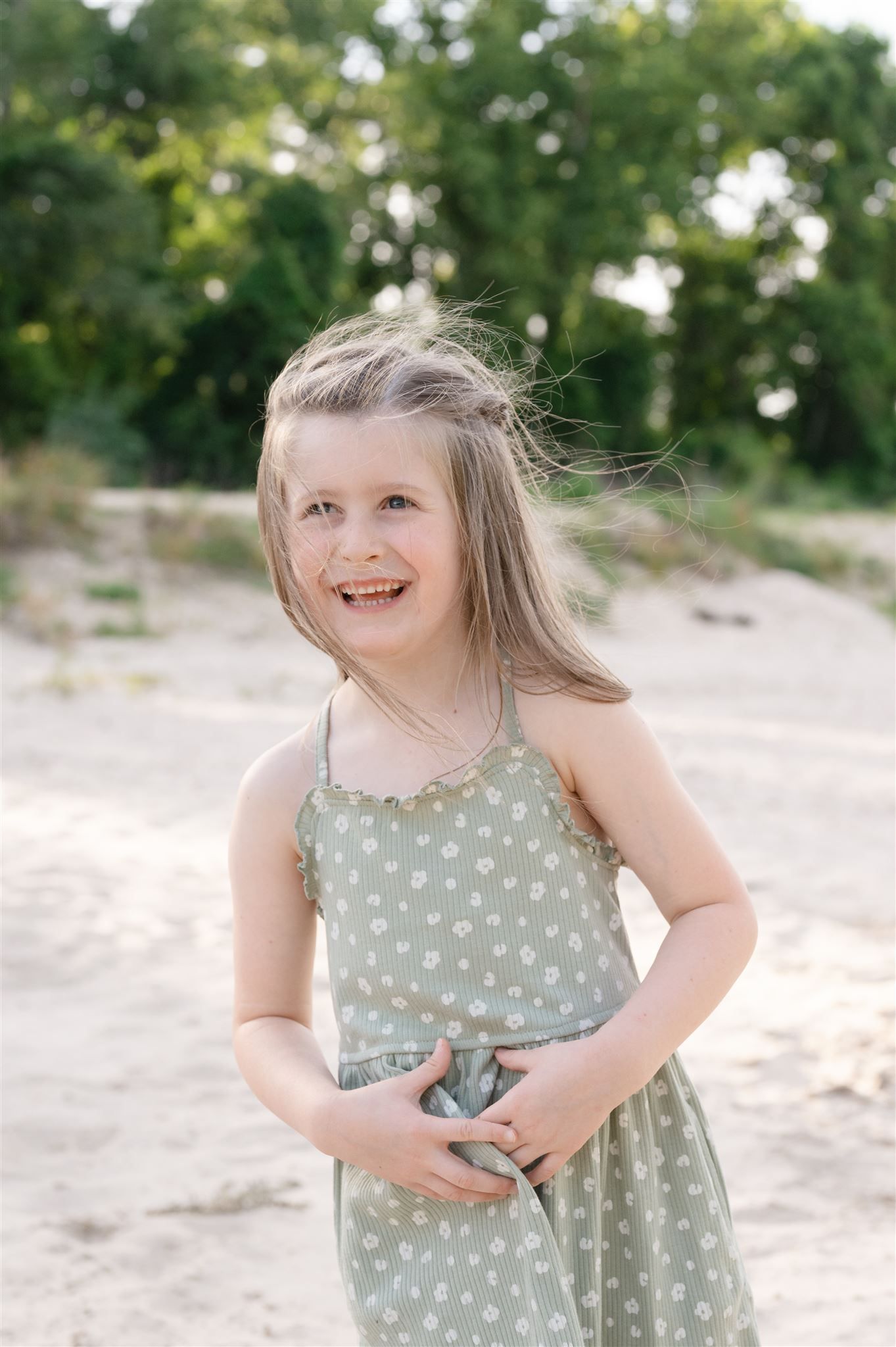 A child wearing a sage green polka dot sundress stands on a sandy beach smiling on a sunny summer day.