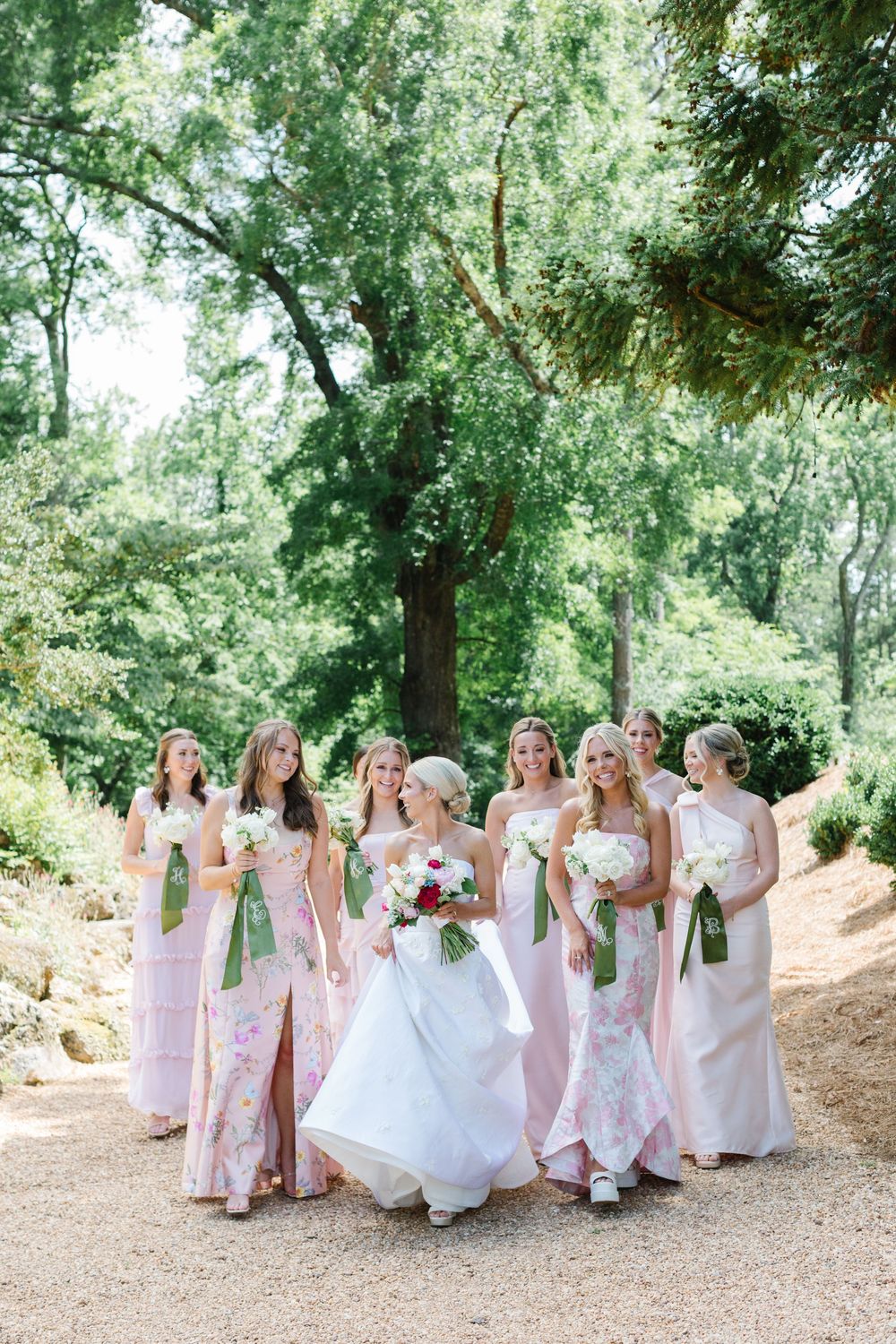 Group of bridesmaids in blush pink gowns with bouquets pose on a wooded path with the bride in white dress.
