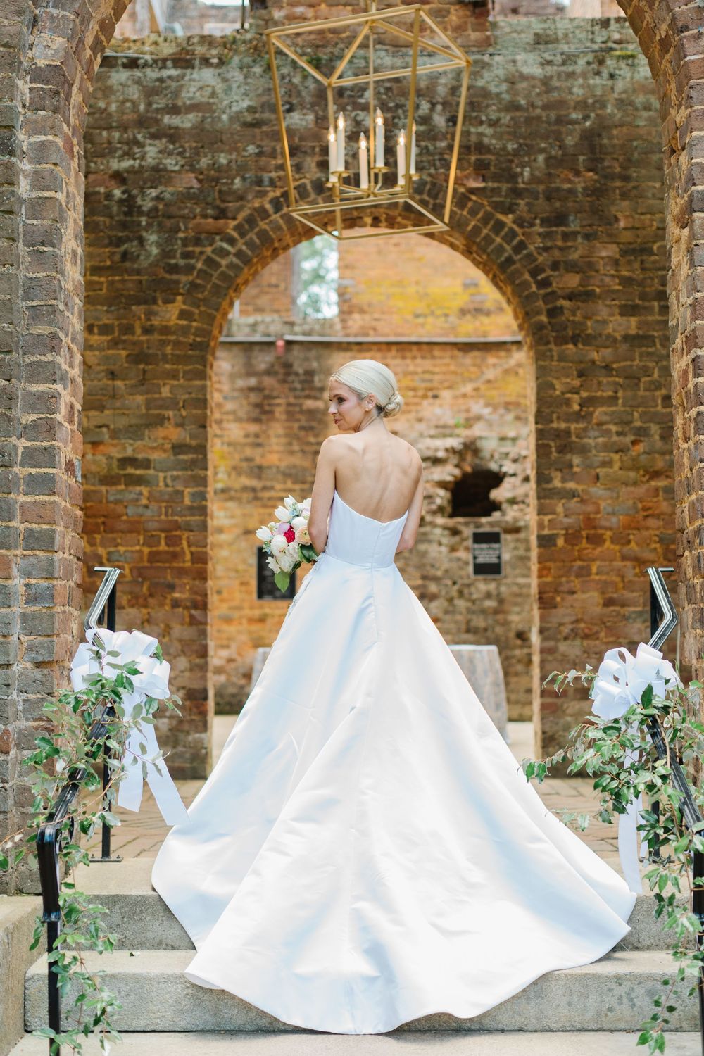 Elegant wedding dress with full skirt and train photographed from behind at historic brick archway with floral decorations.