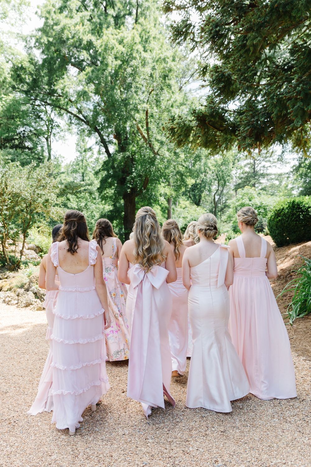 A group of bridesmaids in light pink and white gowns walking down a garden path surrounded by greenery.