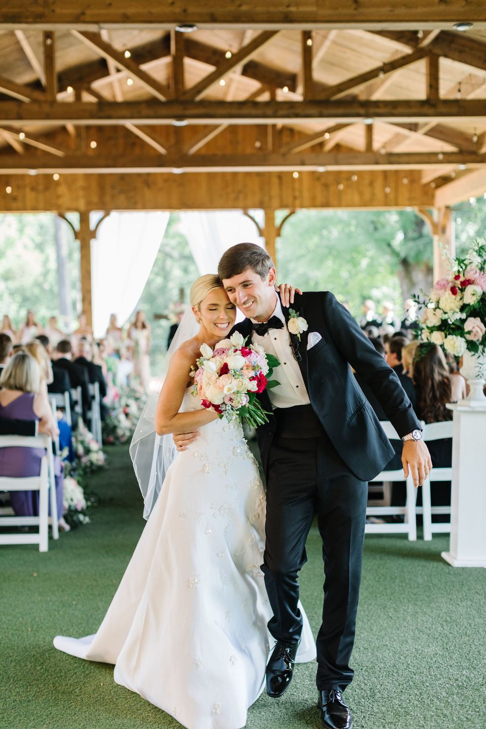 A couple shares an intimate moment at their wedding ceremony under a rustic wooden pavilion with floral decorations.