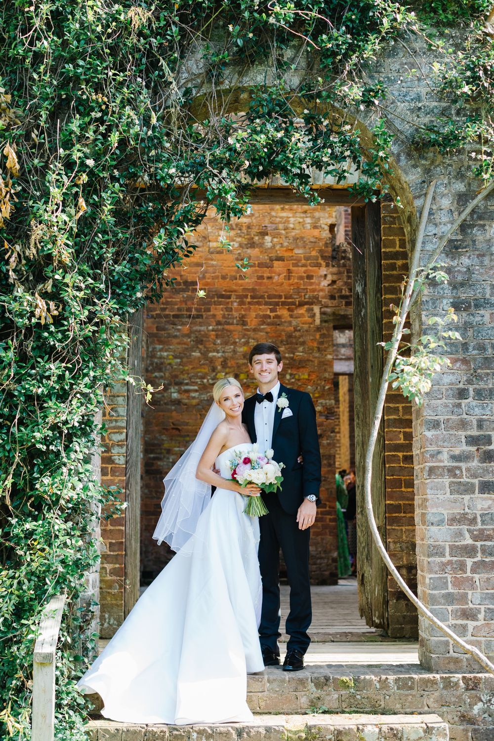 A romantic wedding couple stands in a brick archway covered with lush green ivy at a rustic garden venue.