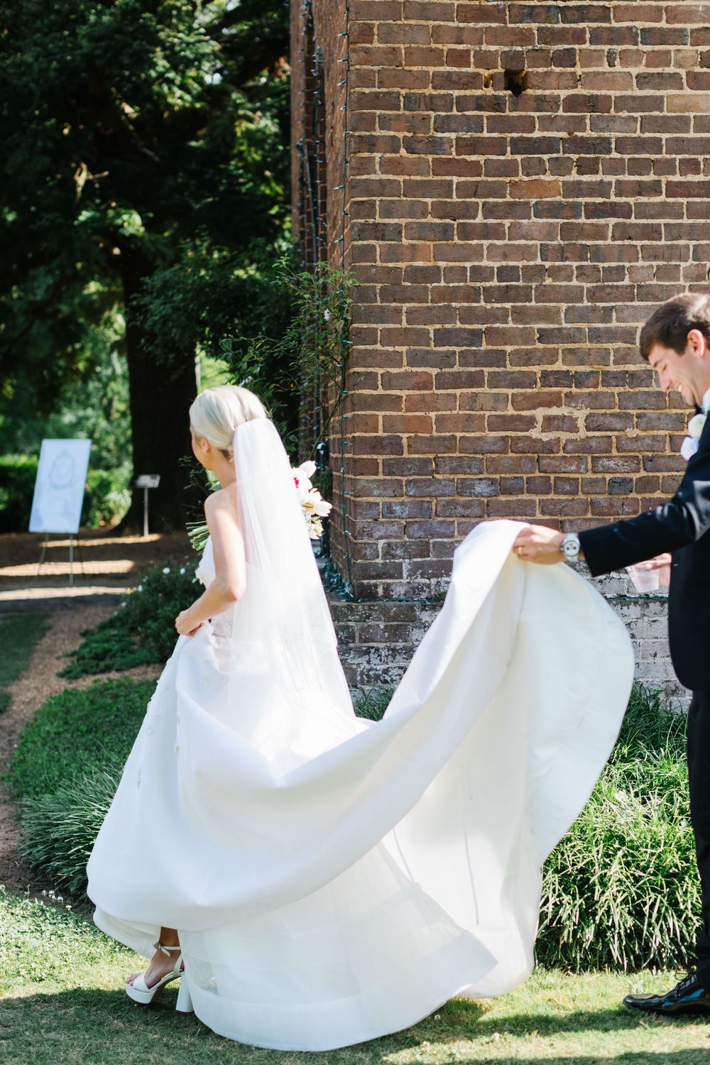 A wedding dress flows elegantly in the breeze as a couple poses against a rustic brick wall outdoors.