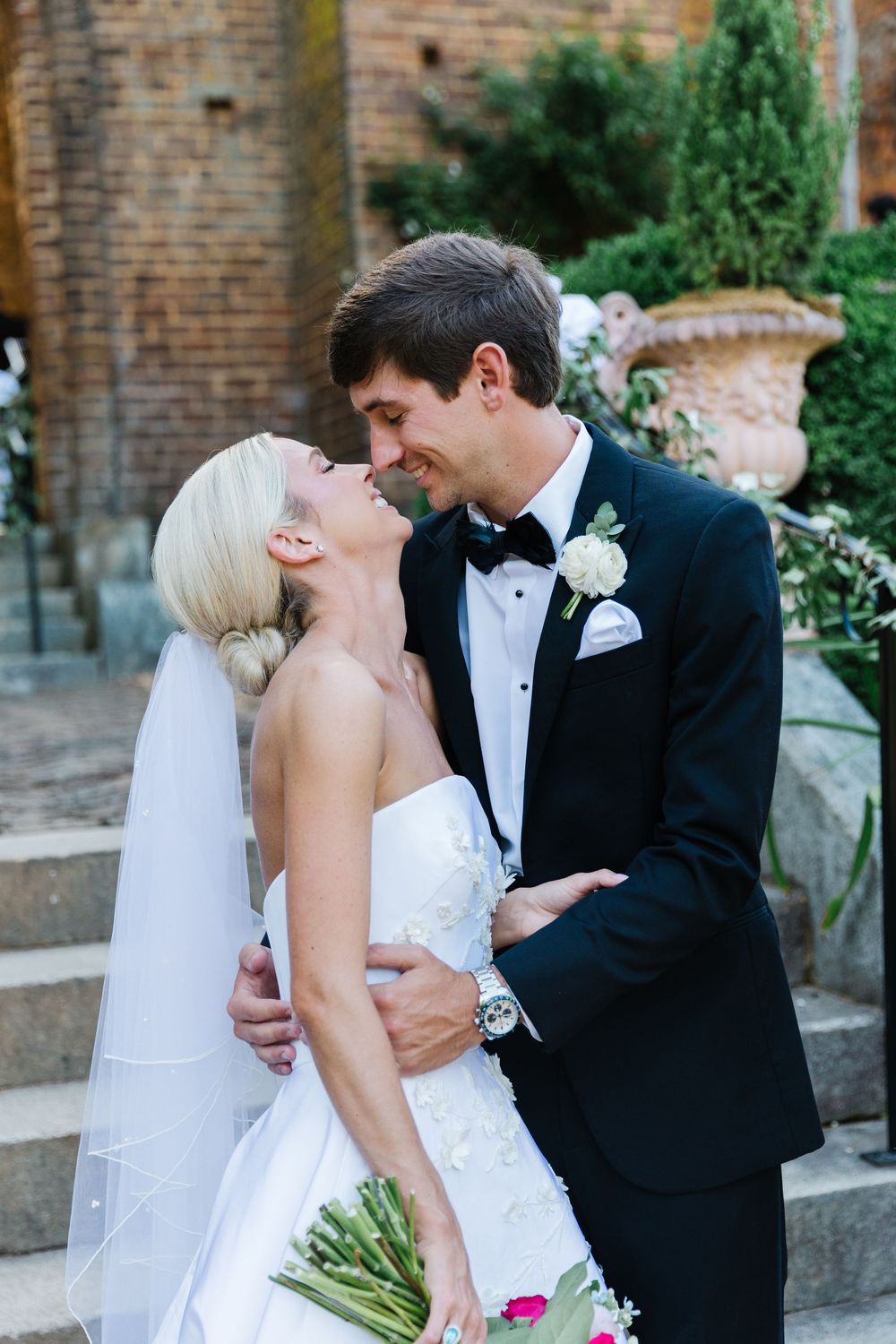 A romantic moment between bride and groom on stone steps with a brick backdrop, featuring a bridal bouquet and formal attire.