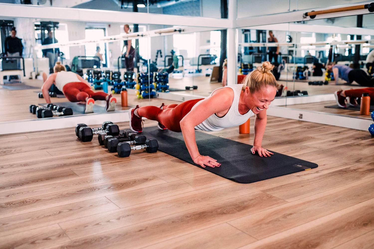 Someone performs a plank exercise on a black yoga mat in a modern gym with dumbbells and exercise equipment.