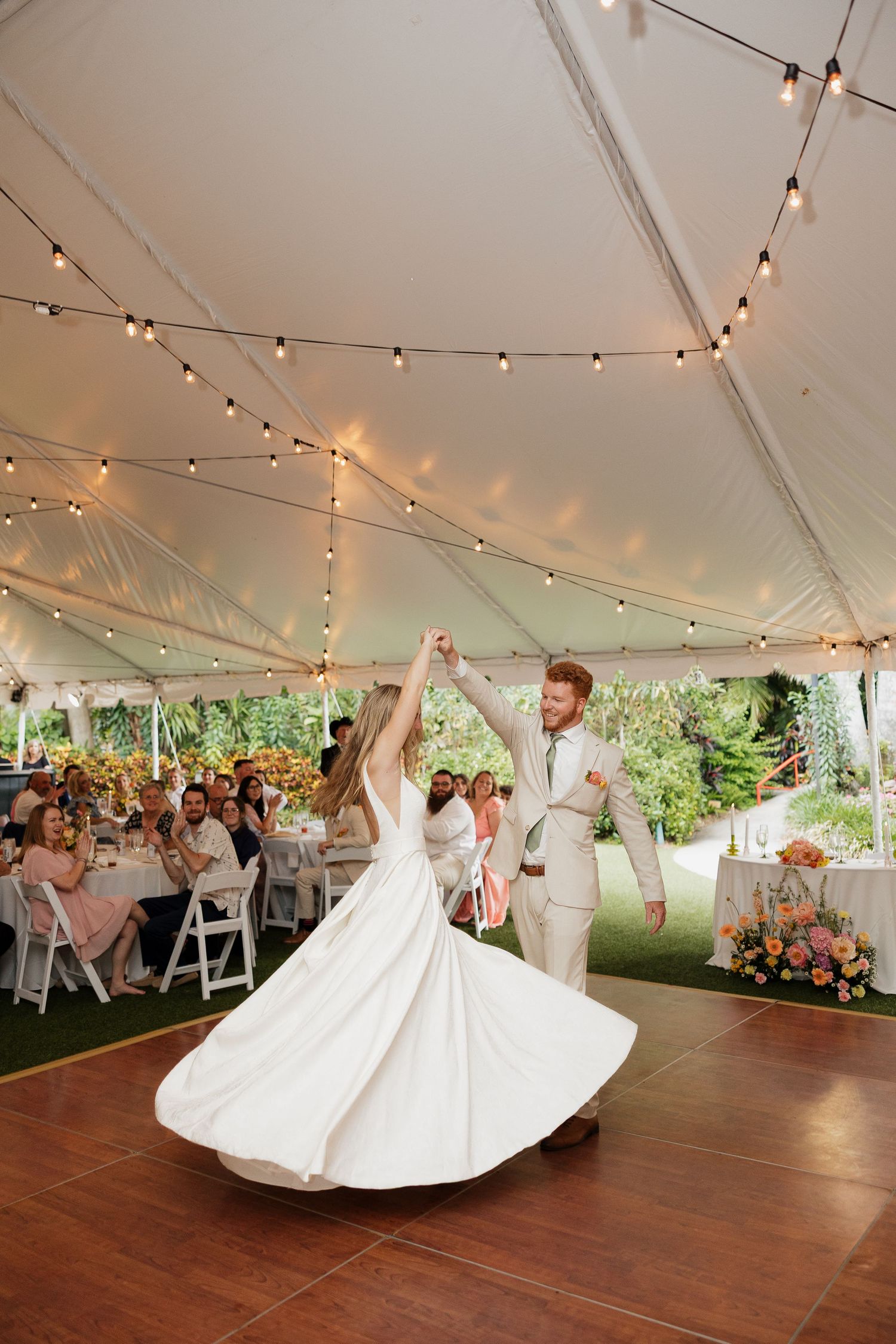 A couple dances their first dance under a tent with string lights at an outdoor wedding reception.