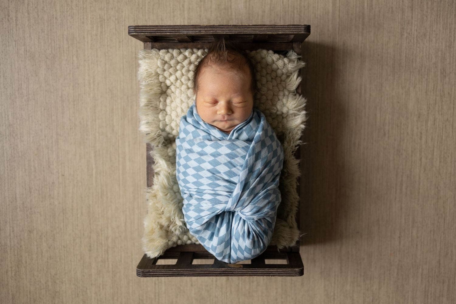 A sleeping newborn wrapped in soft pink and white fabric lies in a tiny baby bed while hands form a heart shape above.