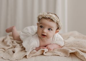Baby in vintage lace dress and bonnet lying on cream colored fabric in soft natural light.