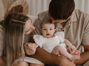 A baby in a white dress sits on a lap while someone reads from a book in warm, neutral tones.