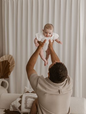 Parent playfully lifts baby in white onesie against neutral curtain background in series of connected photos.