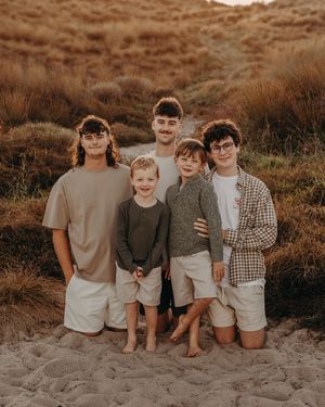 Family standing together on sandy beach at sunset wearing casual coordinating neutral-toned outfits. Photographed on Papamoa beach Tauranga