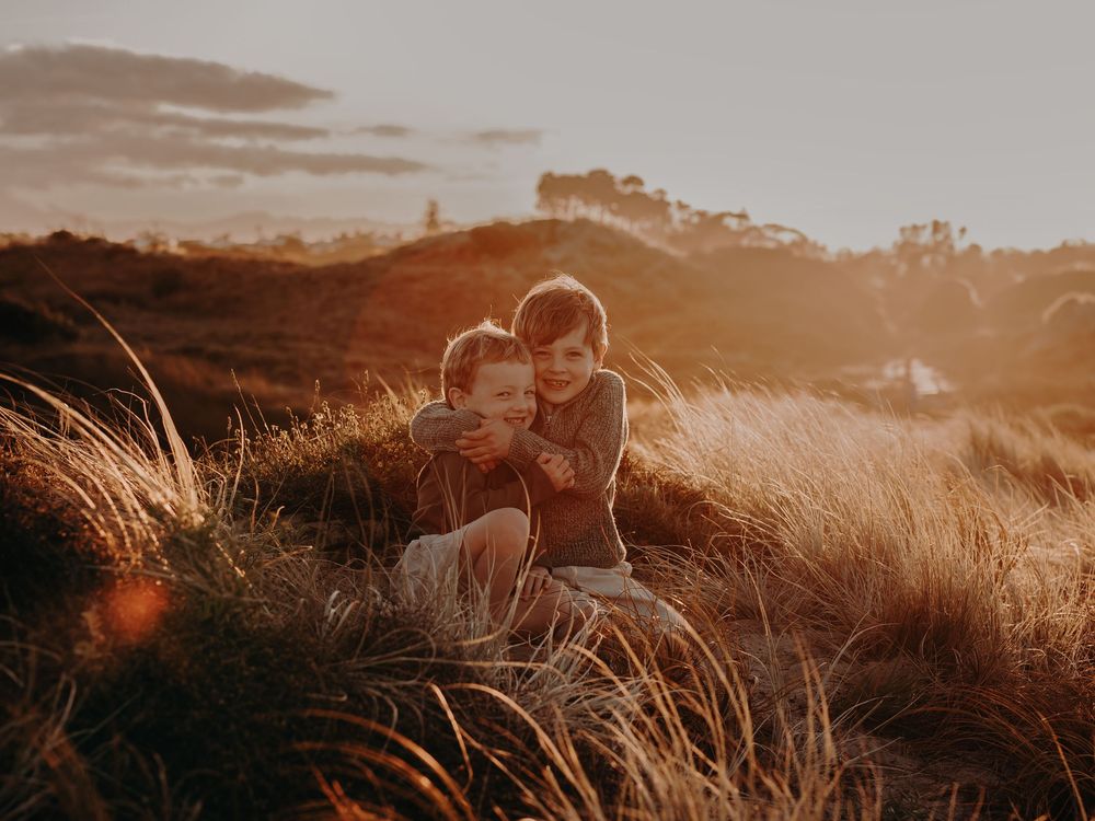Brothers sit together in tall grass during golden sunset, creating a warm silhouetted moment, Photographed on Papamoa beach Tauranga