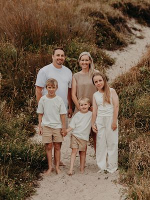 A family of four poses together on a sandy trail surrounded by brown grasses and coastal vegetation.