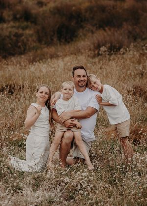 A family wearing white and brown outfits sits together in a golden field during sunset for a candid photo session.