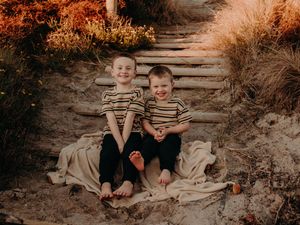 Two children wearing matching striped shirts sit together on rustic outdoor steps surrounded by autumn foliage.