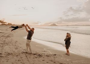 Two people playfully interacting on a sandy beach at sunset with ocean waves and mountains in the background.