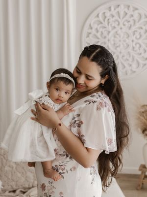 Mother in floral dress holds baby wearing white dress and bow against ornate white backdrop.