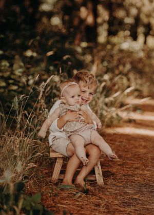 Two young children in light-colored outfits sit together on a reddish dirt path in a warm-toned outdoor setting. Photographed in a Forest