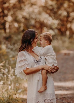 Woman in white lace dress holds and shares tender moment with young child in outdoor autumn setting. Photographed in a Forest