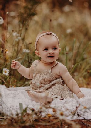 Smiling baby in a beige lace dress sits on white blanket surrounded by wildflowers in a dreamy outdoor setting.
Photographed in a Forest