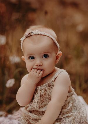 A sweet baby in a lace dress and headband sits outdoors amid autumn colors while holding a hand near their mouth. Photographed in a Forest