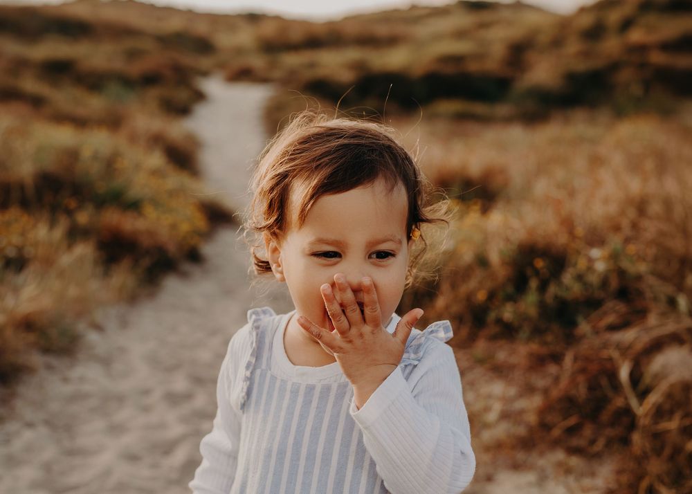 Child in white dress giggles while covering mouth on nature path.