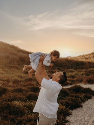 Parent playfully lifting child in white clothing against sunset sky at beach.