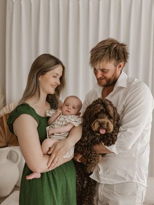A family poses together in a warm indoor setting with their brown curly-haired dog and small baby.