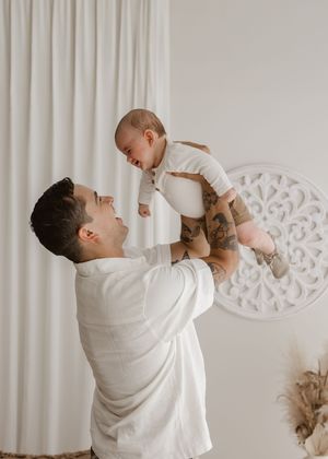 A parent in a white shirt lifts and plays with their baby against a light curtained wall with decorative medallion.