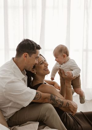 A couple in white and brown clothing shares tender moments with their baby in a bright, sunlit room.