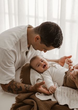 A tender moment between parent and baby lying on white bedding in soft natural light.