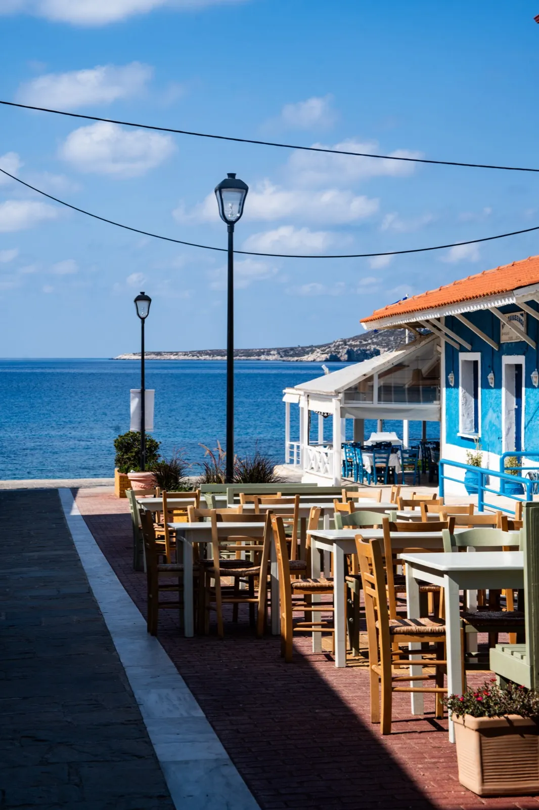 Alignement de chaises en bois et de tables sur une terrasse en bord de mer, baignée de lumière, avec vue dégagée sur l’horizon marin.