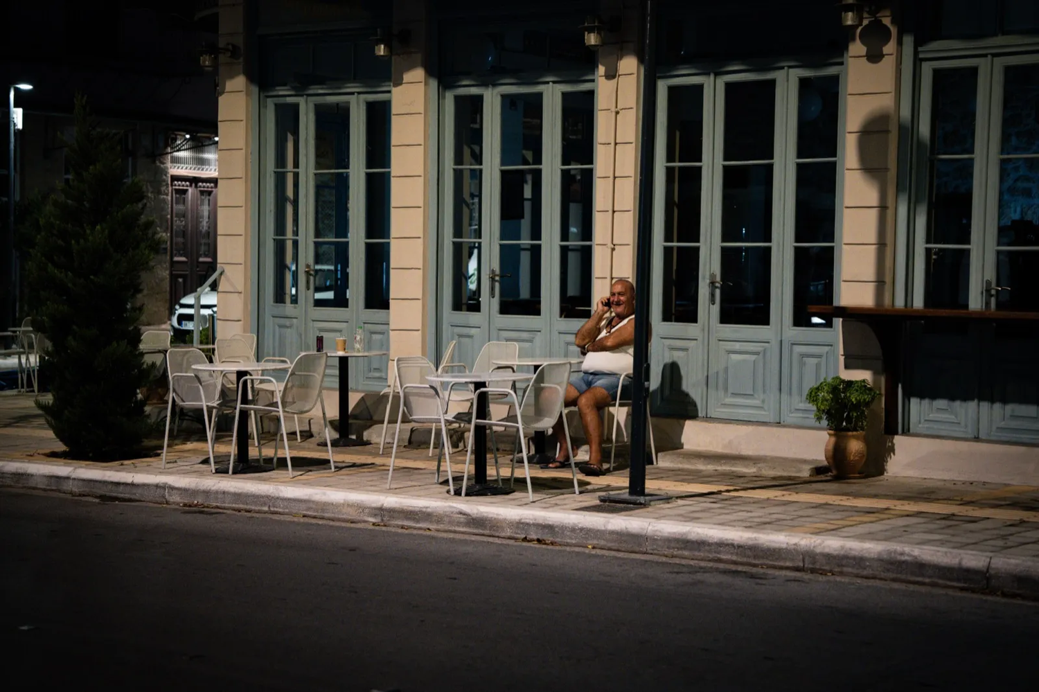 Terrasse de café aux chaises blanches installées devant une façade turquoise ; une personne assise seule, absorbée dans un moment calme.