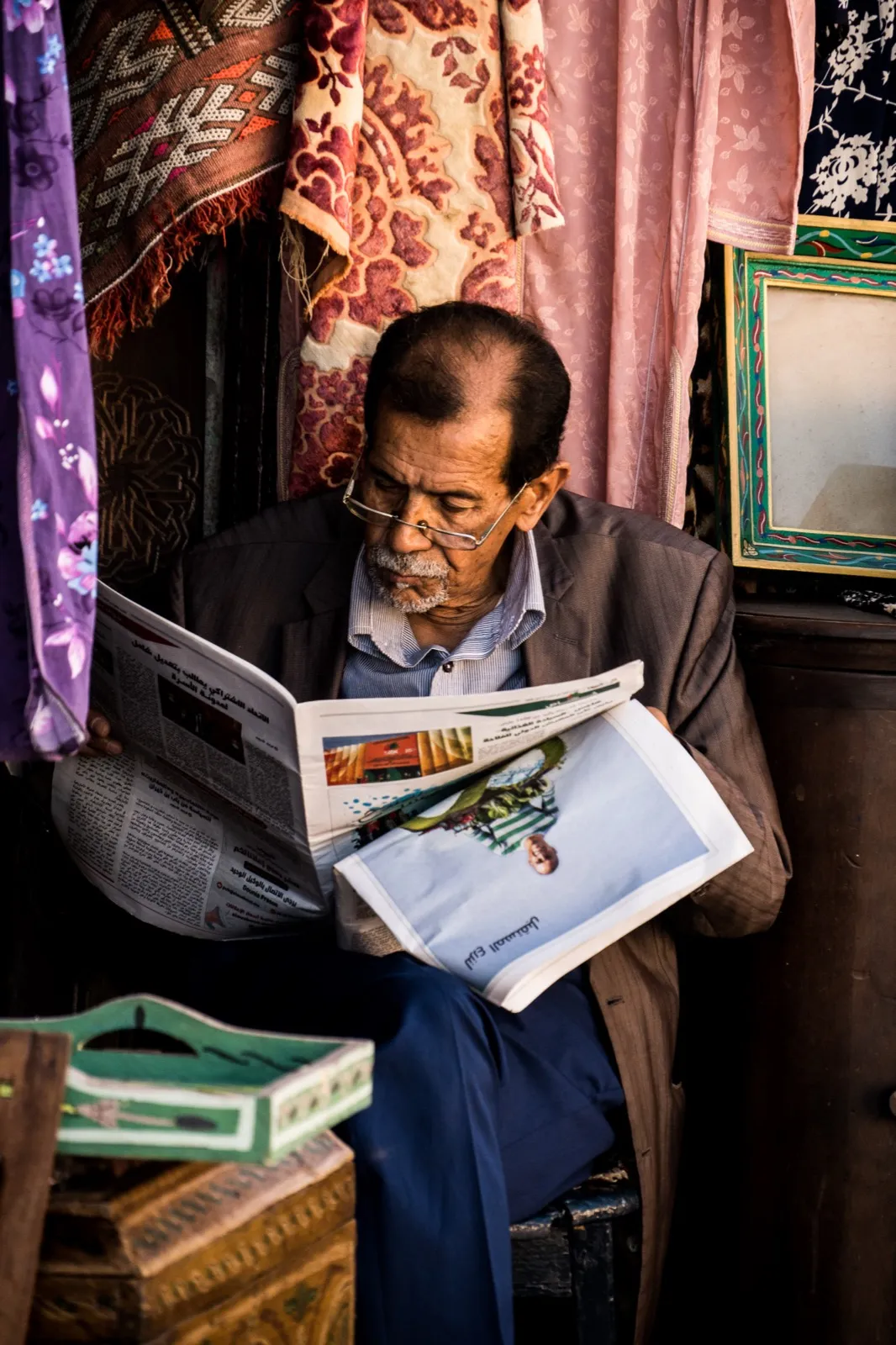 Homme assis dans une boutique ou un intérieur coloré, feuilletant un magazine avec attention, entouré de tissus et d’objets du quotidien.