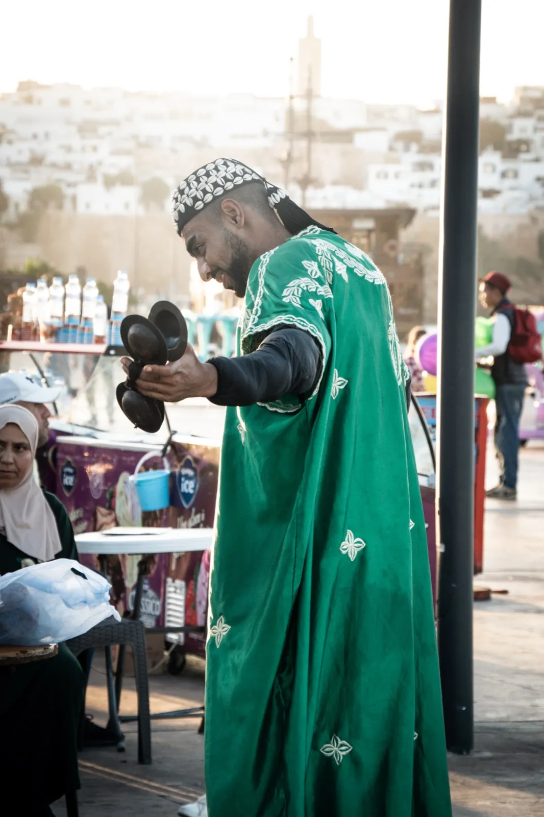 Personne en longue robe verte, entourée de pigeons, nourrissant les oiseaux dans un espace public animé, probablement une place.