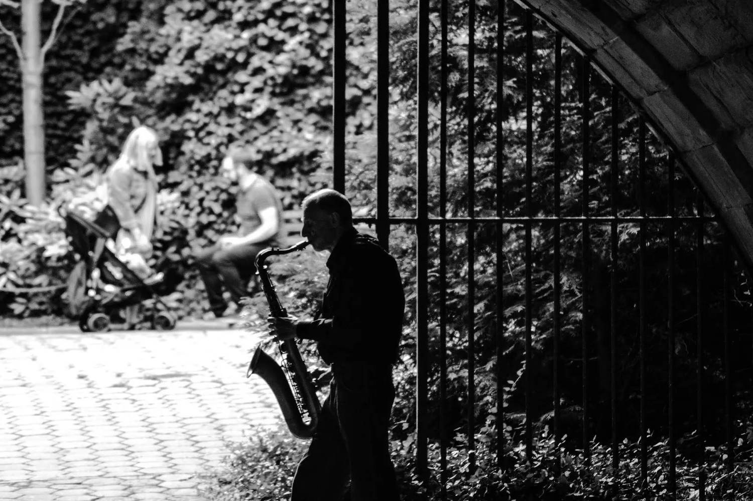 Scène en noir et blanc d’un homme assis à l’ombre d’une arche métallique, regardant un autre passant de l’autre côté de la grille.